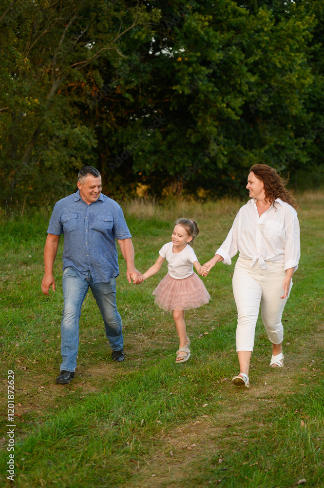Fototapeta premium Family walking together in summer. Mom, dad, daughter walking in meadow holding hands, smiling happily, girl jumping happily. Family, love and concept of parenthood, happy childhood.