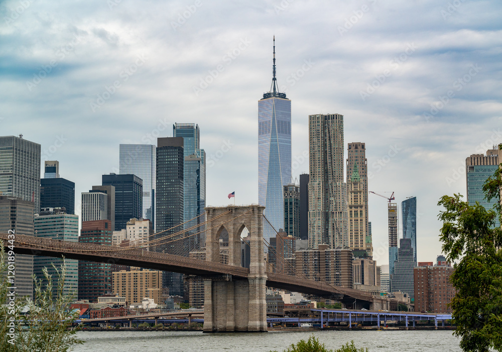 Obraz premium View of the Manhattan skyline with the Brooklyn Bridge and One World Trade Center under a cloudy sky in New York City during late afternoon