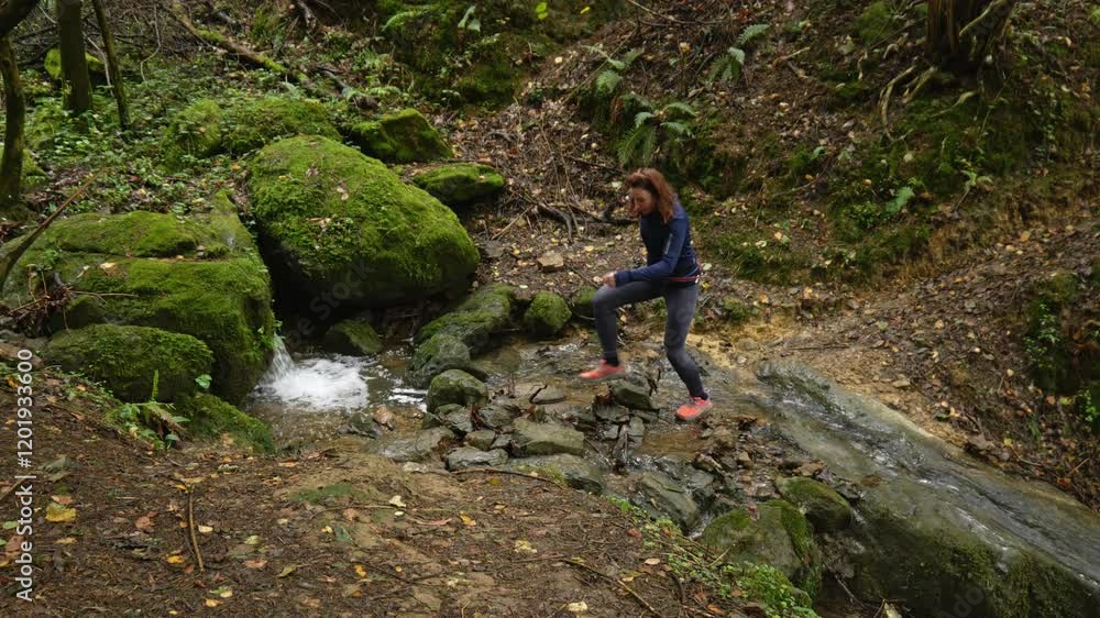 A woman with red hair is walking in the middle of a stream in a forest. There is a small waterfall. A woman with red hair is walking in the middle of a stream in a forest. There is a small waterfall.
