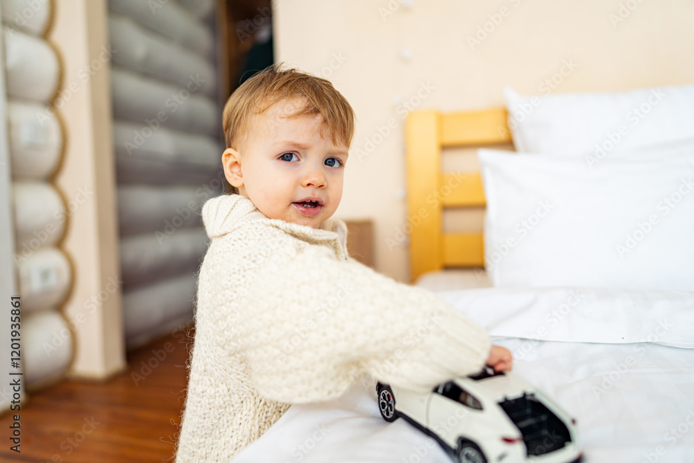 Fototapeta premium Child plays with toy car in cozy bedroom. A young child in a knit sweater engages with a toy car on a bed in a warm, inviting bedroom environment.