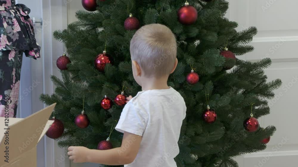 Mother and little boy decorating Christmas tree at home. Happy three year old child helps mom by hanging red ball on christmas tree. Mother and little boy decorating Christmas tree at home. Happy three year old child helps mom by hanging red ball on christmas tree.