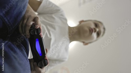 Vertical video of a boy of European appearance in a white tank top and jeans playing a console controlling a red joystick with two hands while looking at the TV, focus on the console from bottom to