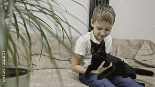 A boy of European appearance with brown hair in a white T-shirt and blue jeans is sitting on a beige sofa and holding a black cat in his hands, says it and smiles at the camera. High quality 4k