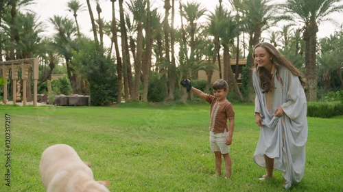 Boy and woman run after two dogs on green lawn surrounded by palm trees in garden of family estate. Woman wears long traditional dress, boy in brown shirt and white shorts. Atmosphere is fill with joy