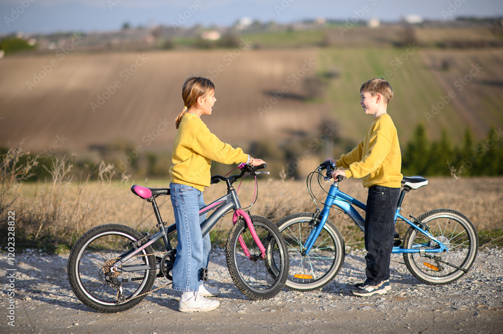 Fototapeta premium A boy and a girl race their bicycles through the countryside at sunset, laughing and chasing each other. The golden light and peaceful surroundings create a joyful, carefree moment of childhood advent