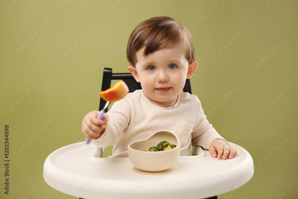 Cute little baby eating healthy food from bowl in high chair on olive background Cute little baby eating healthy food from bowl in high chair on olive background