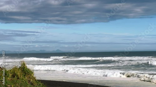 Deserted beach with sea waves washing coast with green grass, in cloudy windy weather. Beginning of storm in ocean