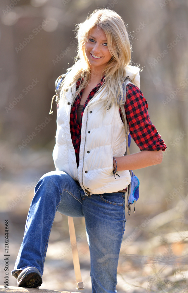 Obraz premium Stunning young blonde model in a a red and black flannel shirt, white down vest, and jeans on hike in woods - climbing over log