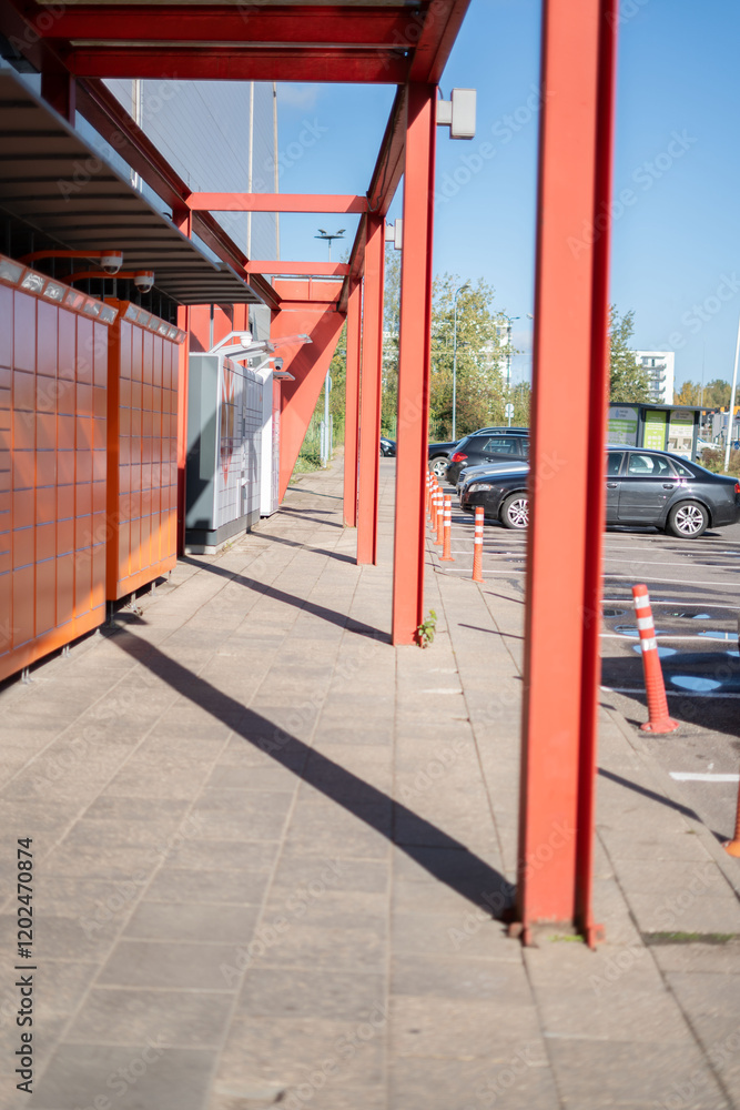 Modern urban delivery lockers in a parking area behind striking red pillars with cars and trees indicating innovative logistics solutions and convenience Modern urban delivery lockers in a parking area behind striking red pillars with cars and trees indicating innovative logistics solutions and convenience