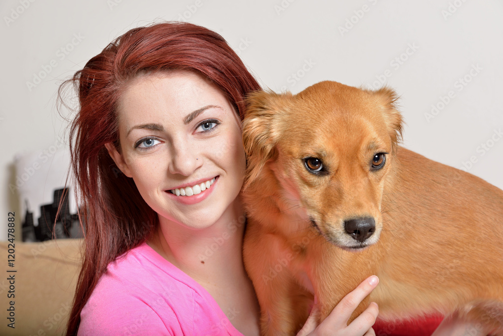 Beautiful young red-haired woman in pink pajamas sits on couch snuggling with adorable dog