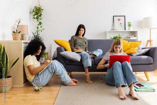 Three female students studying together at home, sitting comfortably on the floor and sofa, engaged in their academic assignments. Female friendship and youth concept photo