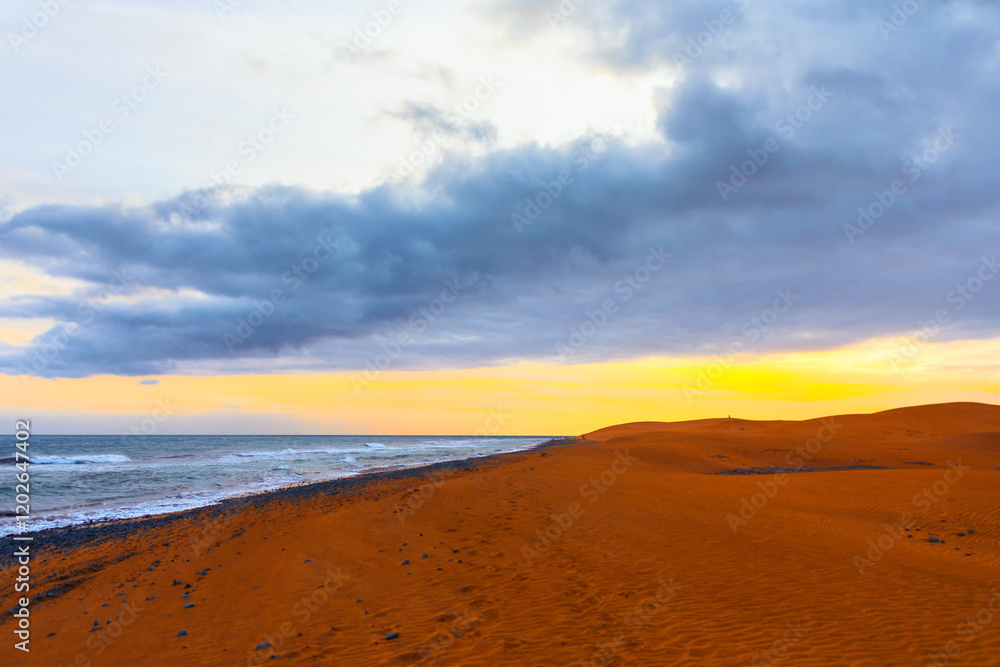 Coastal landscape with a sandy beach extending into distance, bordered by ocean on left and sand dunes on right. Sky is partly cloudy with mix of dark and light clouds, peaceful and picturesque scene