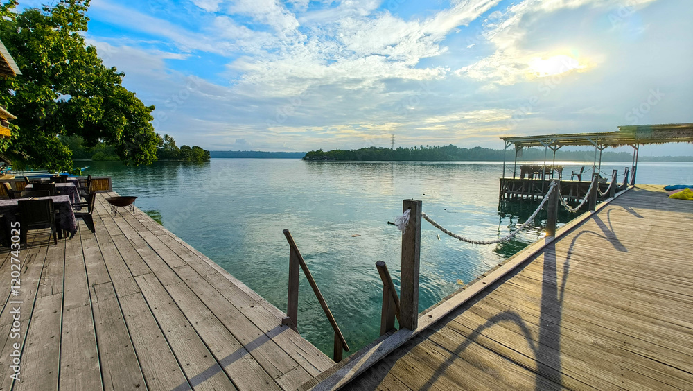 Obraz premium A peaceful waterfront scene with wooden platforms extending over calm waters, surrounded by lush greenery. The serene environment under a partly cloudy sky invites relaxation and reflection