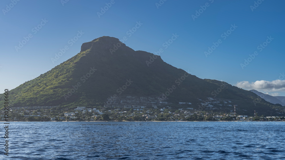 A picturesque mountain against an azure sky and clouds. At the foot and on the slope, urban buildings are visible among the green vegetation. The ocean is in the foreground. Ripples on the blue water A picturesque mountain against an azure sky and clouds. At the foot and on the slope, urban buildings are visible among the green vegetation. The ocean is in the foreground. Ripples on the blue water