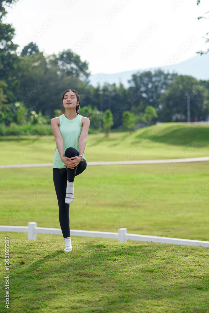 Young asian sportswoman warming up in a park, holding one leg up while closing her eyes, savoring the fresh air on a sunny day surrounded by green grass and trees Young asian sportswoman warming up in a park, holding one leg up while closing her eyes, savoring the fresh air on a sunny day surrounded by green grass and trees
