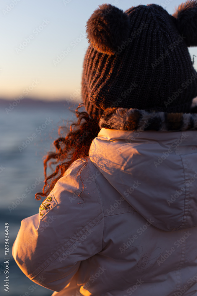 Winter scene: a child observes Lake Garda at sunset, warm light on her white coat.