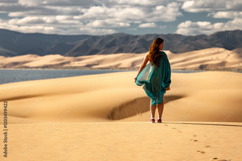 Woman in yellow dress and blue-green scarf is standing at desert dune against blue lake Woman in yellow dress and blue-green scarf is standing at desert dune against blue lake