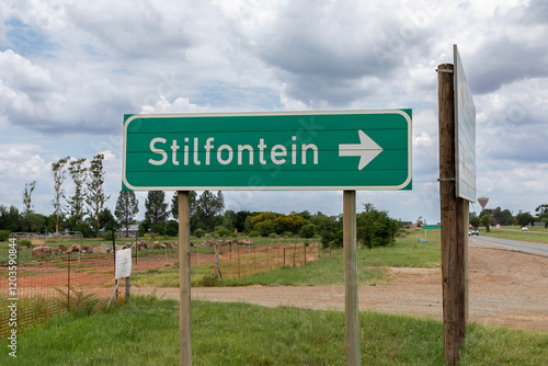 Green Road sign reading Stilfontein with a white arrow pointing to the right.  Stilfontein is a town in South Africa.  
Sign against a cloudy sky. 