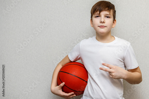 teenage boy in white t-shirt holds orange basketball under arm and shows two fingers