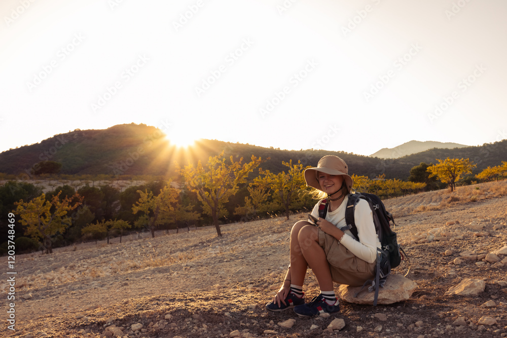 Young female hiker resting on rocky terrain, mountain orchard backdrop glowing with sunset light, smartphone in hand, radiating contentment Young female hiker resting on rocky terrain, mountain orchard backdrop glowing with sunset light, smartphone in hand, radiating contentment