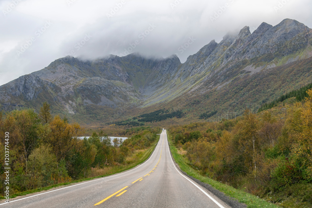 Fototapeta premium Drivers perspective view over European road E10 on Lofoten island chain archipelago in Norway with road disappearing in distance of mountain range with top covered by clouds