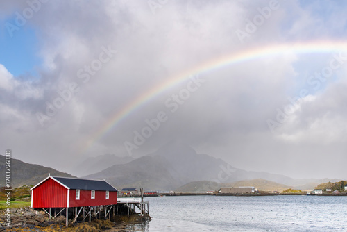 View over a fjord near Reine, Norway on Lofoten island chain archipelago with typical red (fisherman) cabin (rorbu) along shore and dark clouds with colorful rainbow above the landscape photo