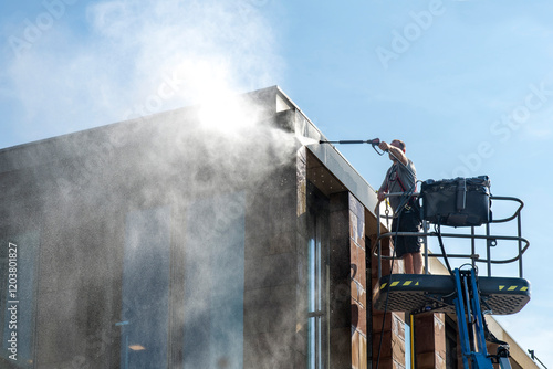 Man standing on aerial work platform or cherry picker with high pressure water gun cleaning the façade of a modern building with water spray against sun covering part of image