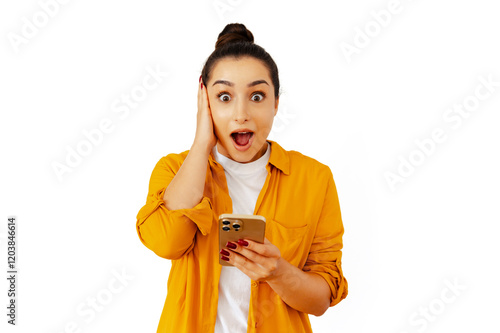 Obraz na plátně Surprised young brunette woman in a yellow shirt holding a smartphone, wide-eyed