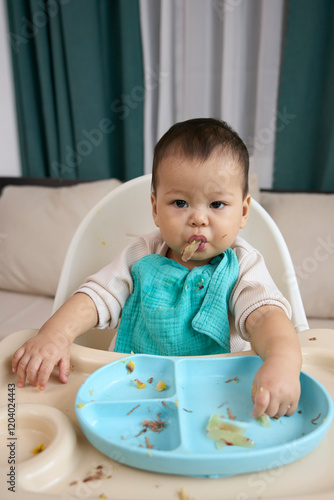 The baby is holding the meat in the mouth while sitting on the dining chair