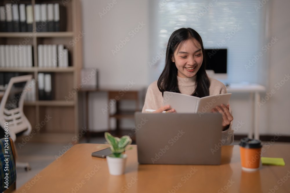 Young Asian business woman smiling and working from home, sitting at a desk and taking notes from a book while using a laptop, with takeaway coffee and smartphone nearby Young Asian business woman smiling and working from home, sitting at a desk and taking notes from a book while using a laptop, with takeaway coffee and smartphone nearby