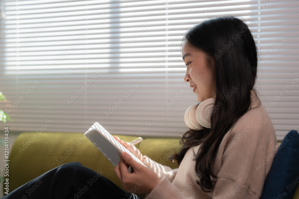 Comfortable young freelancer woman writing notes on notebook while working or studying online and listening to music with headphones, sitting on sofa near window at home Comfortable young freelancer woman writing notes on notebook while working or studying online and listening to music with headphones, sitting on sofa near window at home