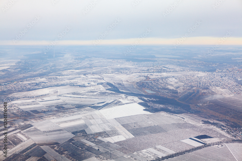 Aerial view of vast landscape covered in snow with patchwork of fields and village visible. Horizon with gradient sky indicating either sunrise or sunset adding serene and picturesque quality to scene