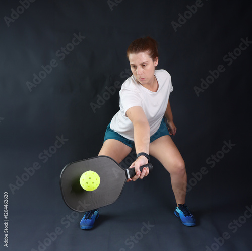 Woman holding a pickleball paddle doing a movement called forehand drop shot with a dark background. Woman in the center of the square photo doing a pickleball dink