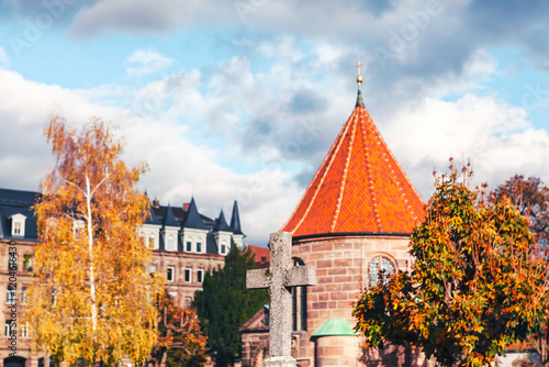 Serene view of Nuremberg Cemetery featuring an old stone cross, autumn trees, and a historic chapel with a red-tiled roof against dramatic sky. Perfect for themes of heritage, history, and tranquility photo