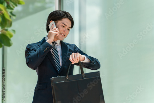 スマホで通話しながら腕時計で時間を確認するスーツ姿の若いアジア人男性ビジネスマン
