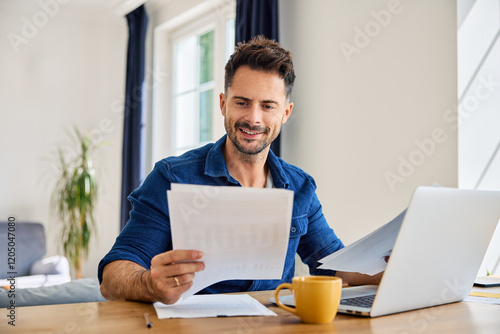 Happy man looking at document while working on laptop from home at living room