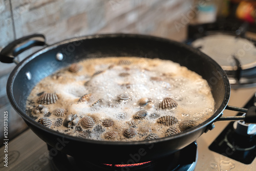 Fresh cockles boiling in a black pan over a gas stove, releasing steam and foam during cooking. photo