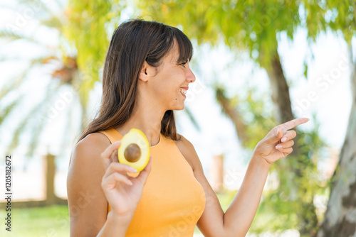 Young woman holding an avocado at outdoors pointing to the side to present a product photo