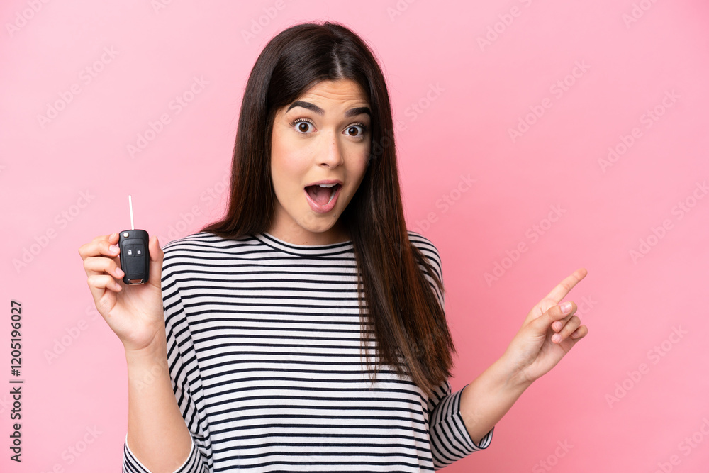 Young Brazilian woman holding car keys isolated on pink background surprised and pointing finger to the side Young Brazilian woman holding car keys isolated on pink background surprised and pointing finger to the side