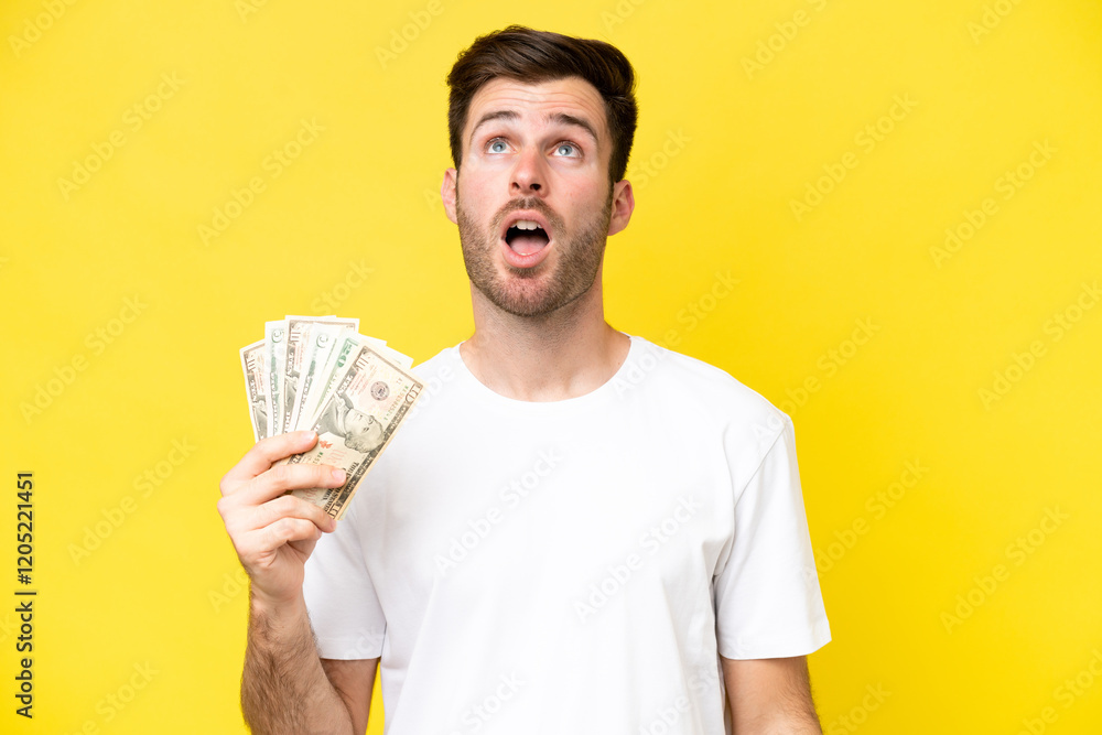 Young caucasian man taking a lot of money isolated on yellow background looking up and with surprised expression Young caucasian man taking a lot of money isolated on yellow background looking up and with surprised expression