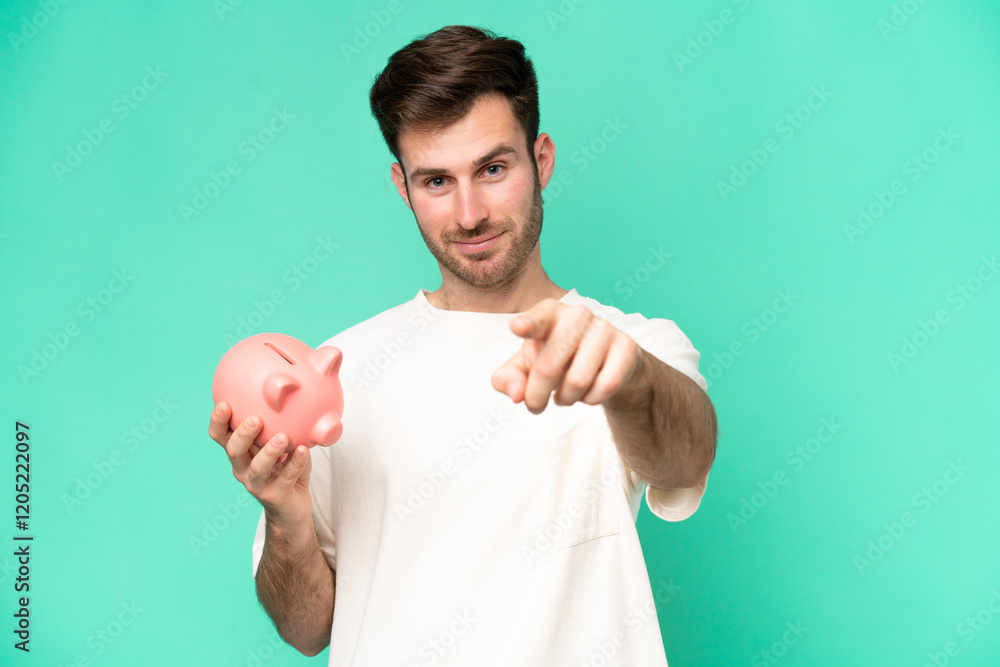Young caucasian man holding piggybank isolated on green background points finger at you with a confident expression Young caucasian man holding piggybank isolated on green background points finger at you with a confident expression
