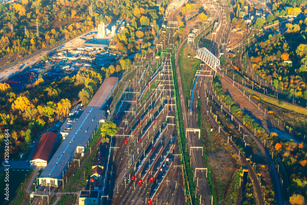 Aerial view of bustling railway hub surrounded by vibrant autumn foliage, showcasing multiple tracks, trains, and industrial buildings. Perfect for themes of transportation, logistics, infrastructure