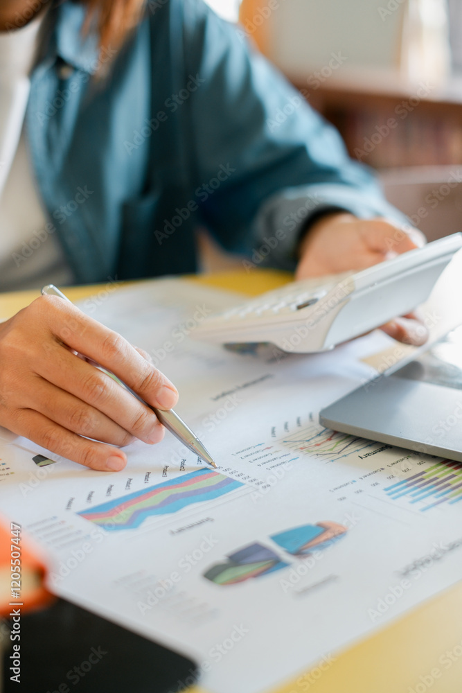 Close-up of a person using a calculator and reviewing financial charts and graphs on paper in an office environment. Close-up of a person using a calculator and reviewing financial charts and graphs on paper in an office environment.