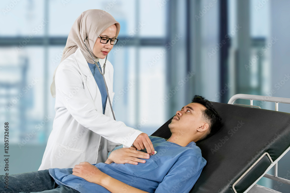 Obraz premium Female Doctor Wearing Hijab Examining a Male Patient with a Stethoscope in a Hospital Bed