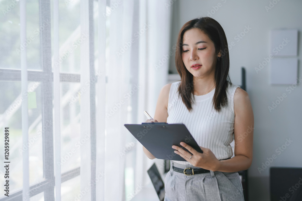 Focused businesswoman reviewing documents and taking notes on clipboard in bright office space