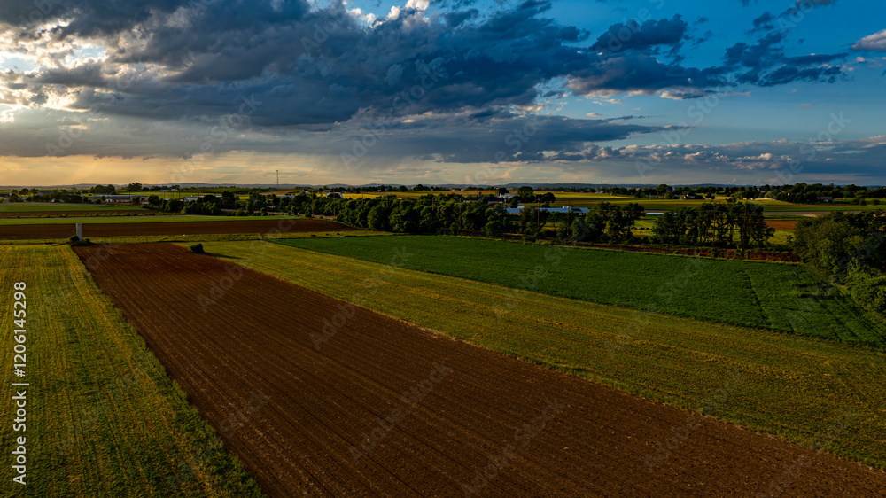Obraz premium As the sun sets in a vibrant sky, vast agricultural fields stretch across the landscape, dotted with trees and farm structures, creating a serene and picturesque rural scene.