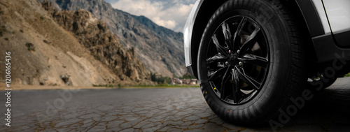 A black tire on a white car is shown close up on a rocky road with mountains in the background.