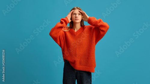 Young teen girl in warm sweater holding her temple, stuffing from a headache and stress on blue studio background. Concept of illness, medicine, treatment, health care photo