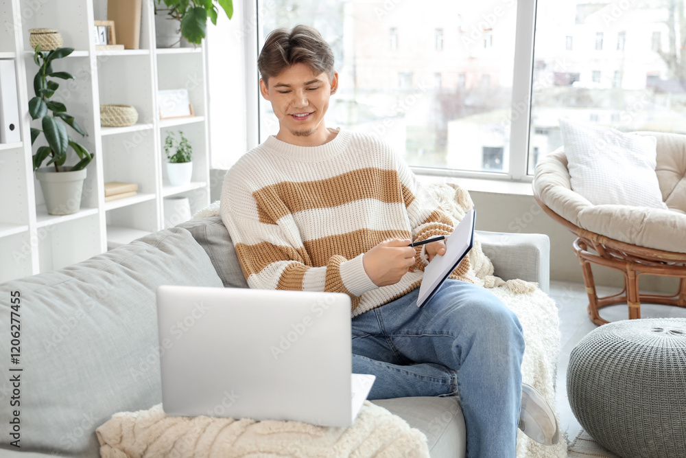 Male student taking notes while studying online on sofa at home