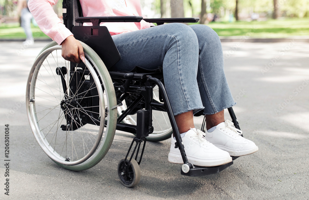 Obraz premium Cropped view of young African American woman using wheelchair outdoors, closeup. Unrecognizable millennial black female with physical impairment enjoying walk in city park